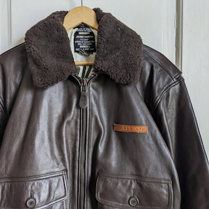 May include: A brown leather jacket with a shearling collar, two front pockets, and a zip closure. The jacket has a label that reads "AVIREX LTD". The jacket is hanging on a wooden hanger.