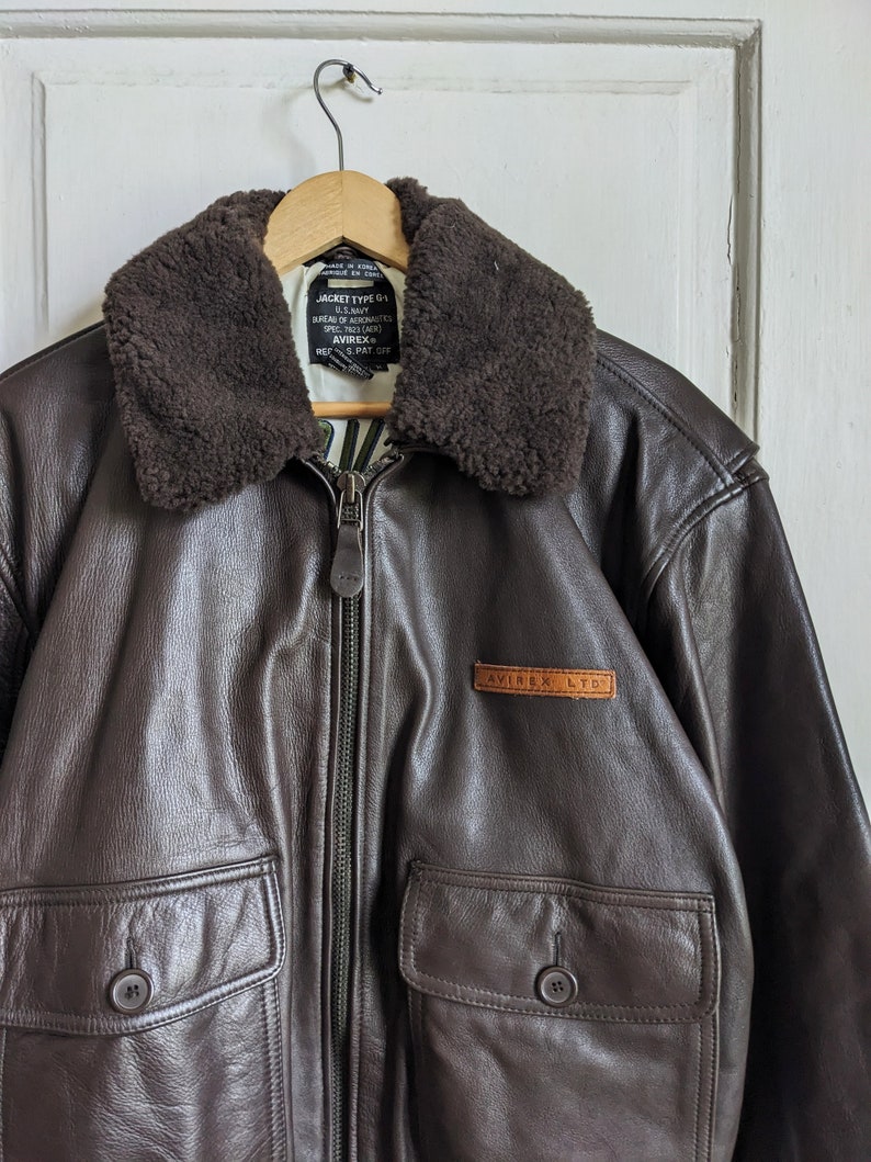 May include: A brown leather jacket with a shearling collar, two front pockets, and a zip closure. The jacket has a label that reads "AVIREX LTD". The jacket is hanging on a wooden hanger.