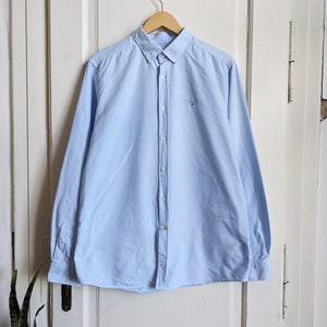 May include: A light blue button-down shirt with long sleeves, hanging on a wooden hanger. The shirt has a small embroidered logo on the chest and a classic collar. The shirt is made of a soft, woven fabric.