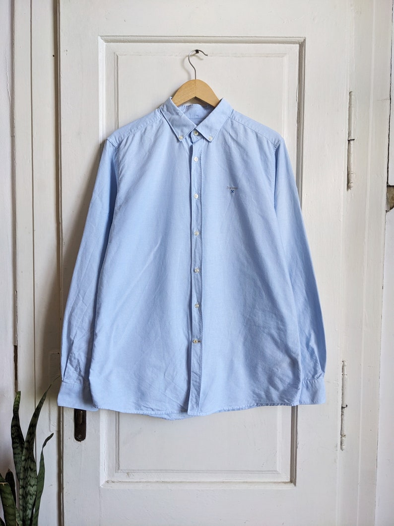 May include: A light blue button-down shirt with long sleeves, hanging on a wooden hanger. The shirt has a small embroidered logo on the chest and a classic collar. The shirt is made of a soft, woven fabric.