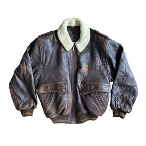 May include: A brown leather bomber jacket with a cream shearling collar. The jacket features two front pockets, a zip closure, and an embroidered design on the chest. Ribbed cuffs and waistband complete the design.