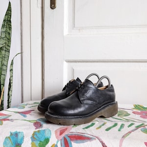 May include: Black leather lace-up shoes with a thick sole. The shoes are sitting on a white tablecloth with a floral pattern.