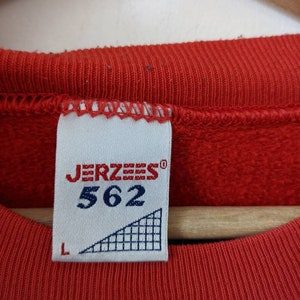 May include: A red sweatshirt with a white tag that says "JERZEES" and "562" in black. The tag also has a white triangle with a black grid pattern and a size "L" label.
