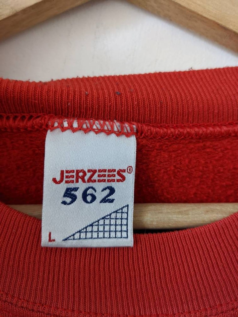 May include: A red sweatshirt with a white tag that says "JERZEES" and "562" in black. The tag also has a white triangle with a black grid pattern and a size "L" label.