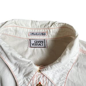 May include: A white button-down shirt featuring contrasting orange stitching along the collar and placket. The shirt includes a gold-toned button and two interior labels, one reading "Made in Italy" and the other "GIANNI VERSACE" in black text.