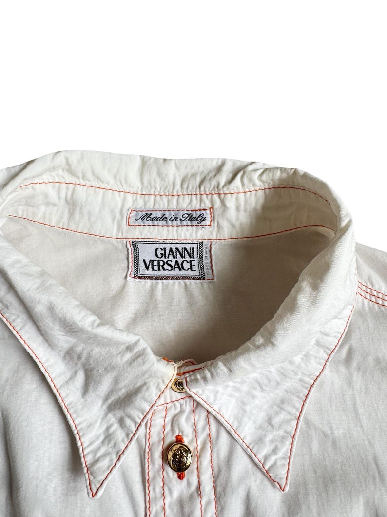 May include: A white button-down shirt featuring contrasting orange stitching along the collar and placket. The shirt includes a gold-toned button and two interior labels, one reading "Made in Italy" and the other "GIANNI VERSACE" in black text.