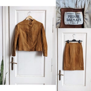 May include: A brown suede jacket and skirt set by Pierre Balmain Paris. The jacket has a stand-up collar, a button closure, and long sleeves. The skirt is a-line and knee-length.