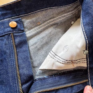 Op de afbeelding: Close-up van een donkerblauwe denim jeans met een gedeeltelijk geopende messing rits. De binnenvoering is zichtbaar, samen met een knoopsluiting en witte stikseldetails. De jeans lijken vintage te zijn.