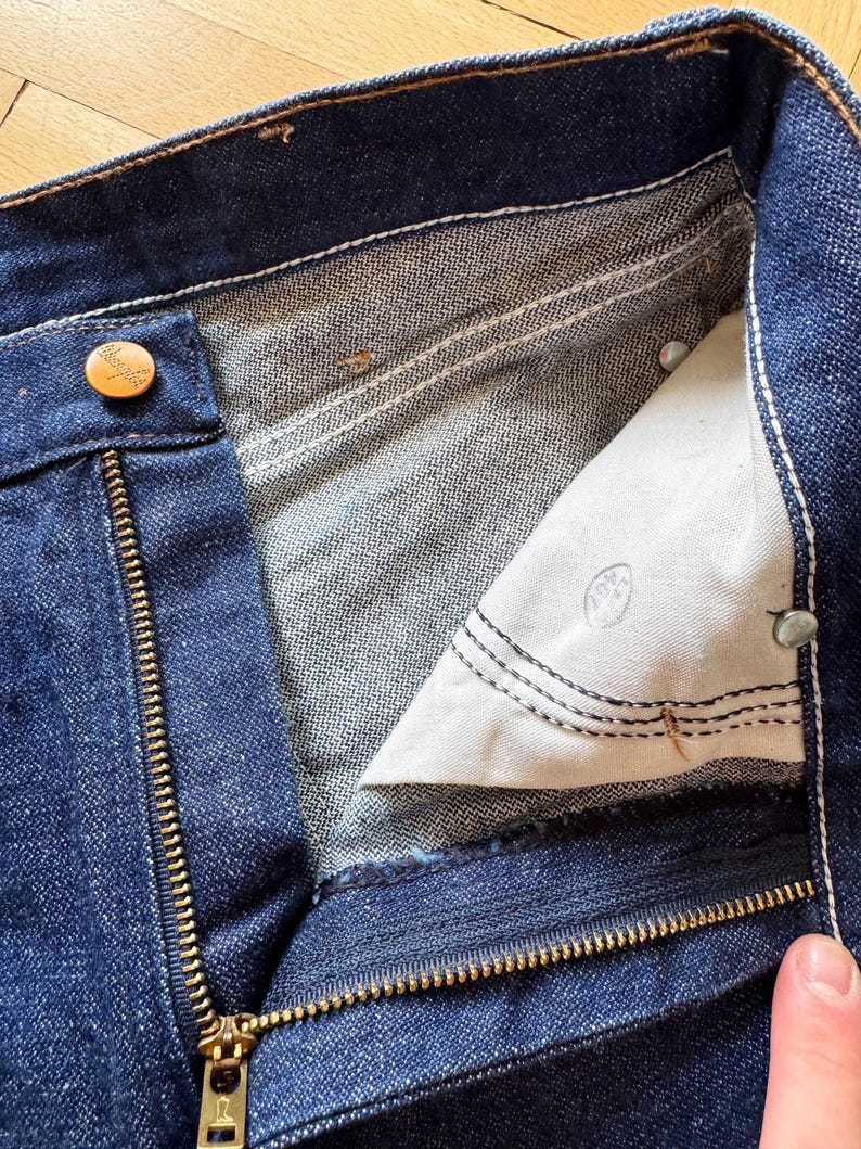 Op de afbeelding: Close-up van een donkerblauwe denim jeans met een gedeeltelijk geopende messing rits. De binnenvoering is zichtbaar, samen met een knoopsluiting en witte stikseldetails. De jeans lijken vintage te zijn.
