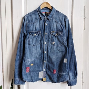Könnte beinhalten: Ein blaues Jeanshemd mit langen Ärmeln, Knopfleiste und mehreren Patches in verschiedenen Farben und Texturen. Das Hemd hat ein Lee-Logo auf der Tasche.