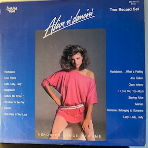 May include: A vintage vinyl record album titled "Alive n' dancin'" featuring a woman in a pink off-the-shoulder top and matching mini-dress. The album cover is blue with song titles listed on the sides. The album is a "Two Record Set".
