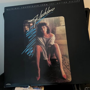 Può includere: Copertina nera dell'album per la colonna sonora originale del film "Flashdance". La copertina presenta una donna con un maglione grigio e tacchi rossi. Il titolo "Flashdance" è in un font stilizzato con accenti blu. L'album è su un giradischi.