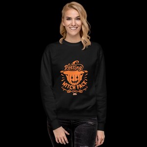 Puede incluir: Una sudadera negra con el texto naranja "Resting Witch Face" y un gráfico de calabaza de Halloween. El diseño presenta una calabaza con un sombrero de bruja y elementos decorativos. Es una prenda cómoda e informal.