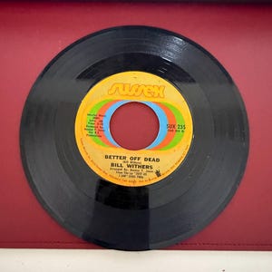 May include: A black vinyl record with a yellow, orange, and blue label. The label reads "BETTER OFF DEAD" by Bill Withers. The record is a 45 RPM single from the Sussex label.