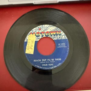 Puede incluir: Un disco de vinilo de 45 RPM vintage de Four Tops, titulado "Reach Out I'll Be There". El disco tiene una superficie negra con una etiqueta azul y blanca con el logotipo de Motown y detalles de la canción. Se adjunta una pegatina amarilla.