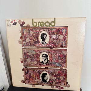 Puede incluir: Portada de álbum de vinilo vintage de la banda Bread. La portada presenta un fondo color crema con el nombre de la banda en verde. Se muestran tres imágenes de billetes de dólar con retratos de los miembros de la banda.