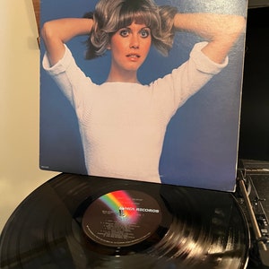 May include: Album cover for Olivia Newton-John's "Don't Stop Believin" featuring a portrait of the artist. The album is on a record player with a black vinyl record. The album cover has a blue background.