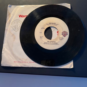Puede incluir: Un disco de vinilo negro de Van Halen, titulado "Girl Gone Bad", se muestra con su funda de papel blanca. La etiqueta del disco presenta el logotipo de Warner Bros. Records y el nombre de la banda. La funda tiene el logotipo y el texto de Warner Bros. Records.