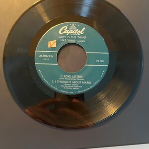 Puede incluir: Un disco de vinilo negro vintage de Nat "King" Cole. La etiqueta del disco es verde azulado y blanca, con el logotipo de Capitol Records y los títulos de las canciones "Love Letters" y "I Thought About Marie". El disco está en buen estado.