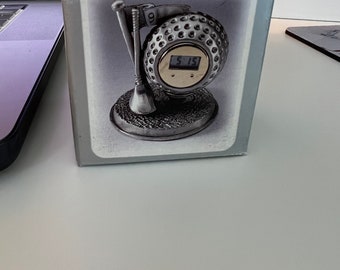 May include: A pewter golf clock in a box. The clock is shaped like a golf ball with a digital display showing 5:15. The clock has a flag and tee design. The box reads "Pewter Golf Clock".