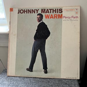 Puede incluir: Portada de álbum de vinilo vintage de "Warm" de Johnny Mathis, con una foto de Mathis con un suéter negro y pantalones oscuros. La portada del álbum tiene el logotipo de Columbia Records y el texto "Percy Faith and His Orchestra."