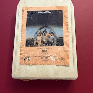 Puede incluir: Un casete de 8 pistas de ABBA Arrival. La etiqueta presenta a los miembros de la banda en una foto de grupo. La cinta está en un cartucho de plástico blanco con el título del álbum y la edición RCA Music Service impresos en la etiqueta. La cinta está sobre una superficie roja.