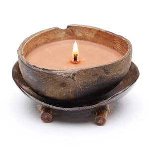 May include: A lit candle in a coconut shell. The candle has a light brown wax and a single wick with a flame. The coconut shell sits on a matching base with three small wooden feet. The background is white.