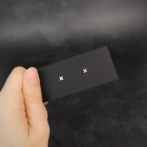 Peut inclure: Une paire de boucles d'oreilles clous argentées en forme de croix, présentées sur une carte noire. Les boucles d'oreilles sont petites et minimalistes, avec un design simple et épuré. Le fond est un dégradé sombre et flou.