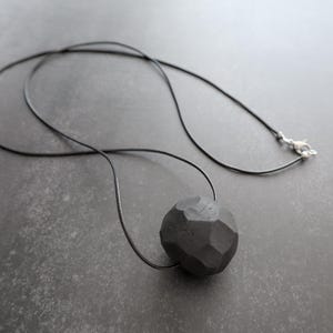 Peut inclure: Un collier noir avec un pendentif géométrique facetté. Le pendentif gris foncé semble être fait de pierre ou d'un matériau similaire. Le collier a un cordon noir fin et un fermoir argenté. Le pendentif mesure environ 3 cm de diamètre.