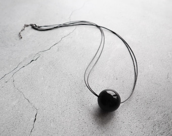 May include: A necklace featuring a single, smooth, spherical black bead suspended on a multi-strand black cord. The cord includes a metallic clasp and an adjustable chain extension at the end, providing a secure and customizable fit for the wearer.