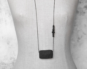 May include: A minimalist black necklace with a rectangular pendant and a series of small beads. The necklace is displayed on a mannequin. The pendant has a textured surface. The necklace is a simple, elegant accessory.