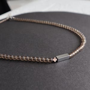 May include: A delicate necklace featuring small, round, smoky quartz beads. A silver-toned, rectangular bar with a small, pink cross accentuates the centre of the necklace. The necklace is displayed on a dark grey surface.