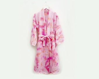 May include: A pink tie-dye robe with a matching belt hangs on a white hanger. The robe features a waffle-weave texture and a vibrant pattern of pink and orange hues. The robe is a comfortable garment for lounging.