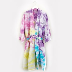 May include: A tie-dye robe with a waffle texture. The robe is white with pink, yellow, blue, and purple tie-dye patterns. The robe has a belt and a pocket on the left side. The robe has the word "Vibes" embroidered on the right side.