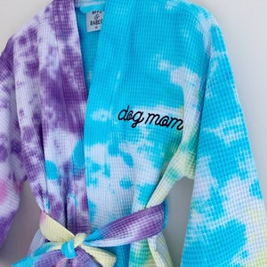 May include: A blue and purple tie-dye waffle weave robe with the text "dog mom" embroidered on the front. The robe has a matching tie belt and is made by the brand "BFF's & Babes".