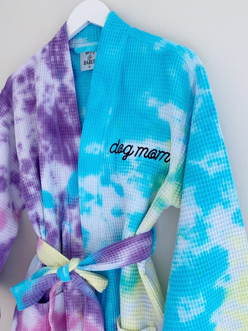May include: A blue and purple tie-dye waffle weave robe with the text "dog mom" embroidered on the front. The robe has a matching tie belt and is made by the brand "BFF's & Babes".