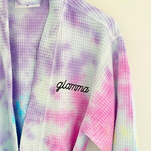 May include: A white waffle weave robe with a tie-dye pattern in shades of purple, pink, blue, and yellow. The robe has the word "glamama" embroidered on the front in black thread.