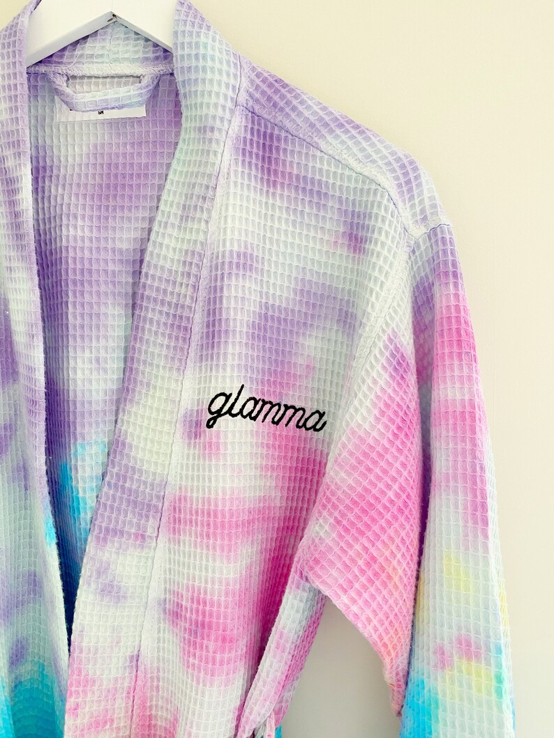 May include: A white waffle weave robe with a tie-dye pattern in shades of purple, pink, blue, and yellow. The robe has the word "glamama" embroidered on the front in black thread.
