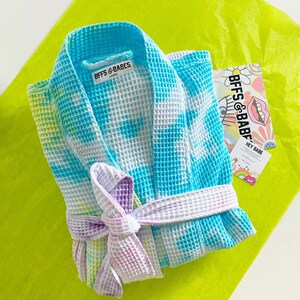 May include: A blue and white tie-dye waffle weave robe with a purple and white tie-dye sash. The robe has a label that says "BFFS & BABES" in black text. The robe is on a bright yellow surface.