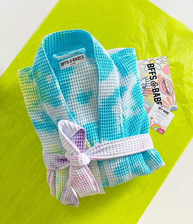 May include: A blue and white tie-dye waffle weave robe with a purple and white tie-dye sash. The robe has a label that says "BFFS & BABES" in black text. The robe is on a bright yellow surface.