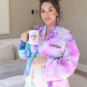 May include: A woman wearing a purple, pink, and blue tie-dye waffle robe with the word "mama" embroidered on the front. She is holding a white mug with a photo of a child on it.