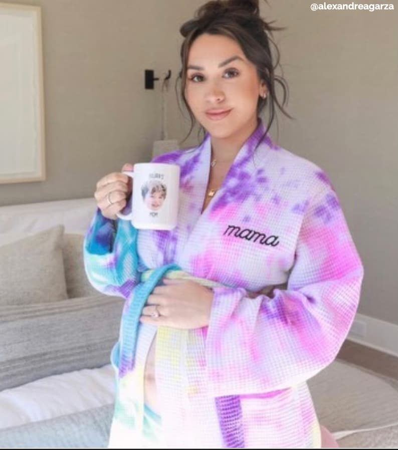 May include: A woman wearing a purple, pink, and blue tie-dye waffle robe with the word "mama" embroidered on the front. She is holding a white mug with a photo of a child on it.