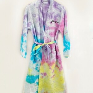 May include: A tie-dye waffle weave robe with a light blue, yellow, and pink color scheme. The robe has a self-tie belt and the word "glama" embroidered on the front.