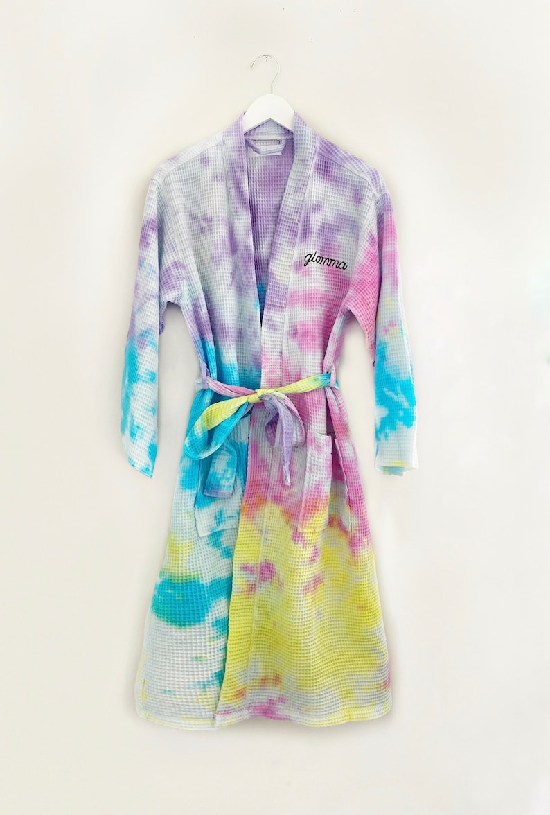 May include: A tie-dye waffle weave robe with a light blue, yellow, and pink color scheme. The robe has a self-tie belt and the word "glama" embroidered on the front.