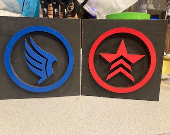 May include: Two wooden signs with black backgrounds and circular cutouts. The left sign features a blue wing symbol in a blue circle. The right sign features a red star with three chevrons in a red circle.