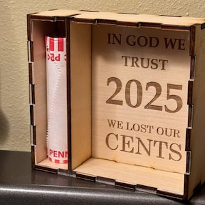 Może przedstawiać: Drewniane pudełko z otworem na rolkę centów. Na pudełku wygrawerowano słowa "IN GOD WE TRUST 2025 WE LOST OUR CENTS". Pudełko jest wykonane z jasnego drewna.