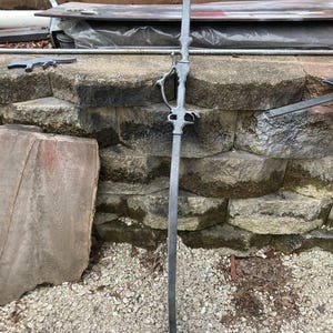 Op de afbeelding: Een zilverkleurig metalen zwaard met een decoratief handvat en een lange, rechte kling. Het zwaard leunt tegen een stenen muur.