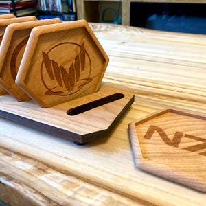 May include: Set of six wooden coasters with a hexagonal shape and a laser-engraved logo. The coasters are arranged in a stack and are sitting on a wooden stand. The logo is a stylized "NZ" with a geometric design.