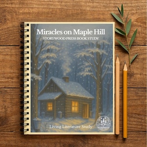 May include: A spiral-bound book titled "Miracles on Maple Hill" with a winter scene illustration. The cover shows a snow-covered cabin with lit windows, surrounded by trees. Two pencils and a sprig of greenery rest on a wooden surface.