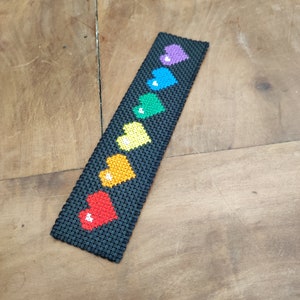 Peut inclure: Un marque-page noir avec un arc-en-ciel de cœurs cousus dessus. Les cœurs sont dans l'ordre de l'arc-en-ciel, avec le rouge en bas et le violet en haut.